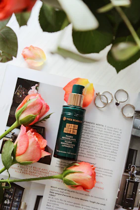 A bottle of Yves Rocher Lifting Vegetal fluid is placed on a magazine with vibrant pink roses around it. Silver rings rest next to the bottle and some rose petals are scattered. The background includes green leaves, adding a natural touch.