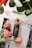 A bottle of Yves Rocher Lifting Vegetal fluid is placed on a magazine with vibrant pink roses around it. Silver rings rest next to the bottle and some rose petals are scattered. The background includes green leaves, adding a natural touch.