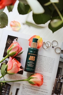 A bottle of Yves Rocher Lifting Vegetal fluid is placed on a magazine with vibrant pink roses around it. Silver rings rest next to the bottle and some rose petals are scattered. The background includes green leaves, adding a natural touch.