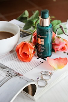 A small bottle of cosmetic serum with a golden cap is placed on an open magazine alongside blooming orange roses. To the side, there's a cup filled with coffee, and a few metallic rings lay next to the flowers. The scene is set on a glossy magazine page, creating an elegant and serene composition.