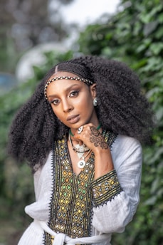 A woman with elaborate braids and voluminous natural hair stands in front of a lush green background. She wears a traditional garment with intricate embroidery featuring geometric patterns in black, yellow, and green. Her hand, adorned with henna, rests near her face, showcasing her jewelry, including a large earring and several necklaces.