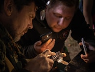 Two individuals are closely examining a small object that appears to be held in a clamp or vise. They are using flashlight apps on their smartphones to illuminate the object.