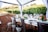 A beautifully set dining table outdoors, surrounded by lush vineyards. The table is adorned with elegant wine glasses, plates, silverware, and vibrant floral arrangements. The chairs are metallic, and there's an overhead canopy with white fabric curtains framing the scene.