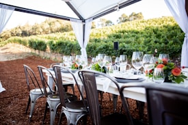 A beautifully set dining table outdoors, surrounded by lush vineyards. The table is adorned with elegant wine glasses, plates, silverware, and vibrant floral arrangements. The chairs are metallic, and there's an overhead canopy with white fabric curtains framing the scene.