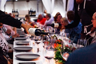 A group of people are seated around a long dining table, which is elegantly set with wine glasses, plates, and floral centerpieces. Two people are pouring red wine from bottles into the glasses. The ambiance is cozy, lit by soft candlelight, creating a warm and inviting atmosphere.