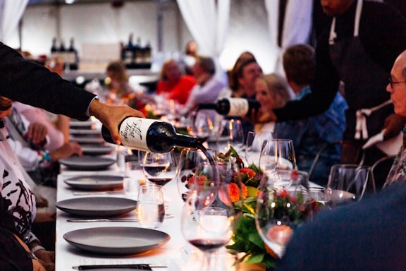 A group of people are seated around a long dining table, which is elegantly set with wine glasses, plates, and floral centerpieces. Two people are pouring red wine from bottles into the glasses. The ambiance is cozy, lit by soft candlelight, creating a warm and inviting atmosphere.