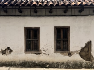 A weathered plaster wall with two wooden-framed windows, both featuring metal bars. The surface shows significant wear, with visible cracks and areas where the plaster has chipped away, revealing underlying materials. Above, the roof is constructed with dark wooden beams supporting traditional clay tiles.