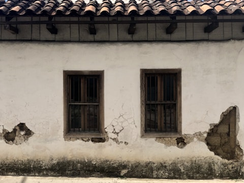 A weathered plaster wall with two wooden-framed windows, both featuring metal bars. The surface shows significant wear, with visible cracks and areas where the plaster has chipped away, revealing underlying materials. Above, the roof is constructed with dark wooden beams supporting traditional clay tiles.