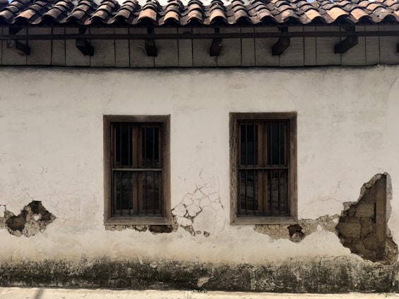 A weathered plaster wall with two wooden-framed windows, both featuring metal bars. The surface shows significant wear, with visible cracks and areas where the plaster has chipped away, revealing underlying materials. Above, the roof is constructed with dark wooden beams supporting traditional clay tiles.