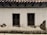 A weathered plaster wall with two wooden-framed windows, both featuring metal bars. The surface shows significant wear, with visible cracks and areas where the plaster has chipped away, revealing underlying materials. Above, the roof is constructed with dark wooden beams supporting traditional clay tiles.