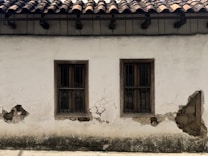 A weathered plaster wall with two wooden-framed windows, both featuring metal bars. The surface shows significant wear, with visible cracks and areas where the plaster has chipped away, revealing underlying materials. Above, the roof is constructed with dark wooden beams supporting traditional clay tiles.