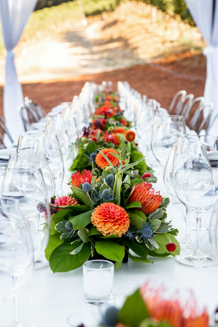 Long banquet table with vibrant orange floral runner set among vineyard rows