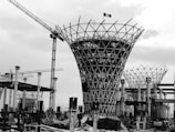 A black and white image of a construction site featuring a large, uniquely shaped structure with a lattice pattern. Several cranes can be seen in the background, along with scaffolding and various construction materials. The sky appears overcast.