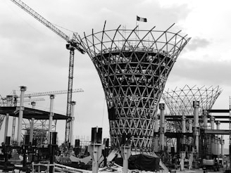 A black and white image of a construction site featuring a large, uniquely shaped structure with a lattice pattern. Several cranes can be seen in the background, along with scaffolding and various construction materials. The sky appears overcast.