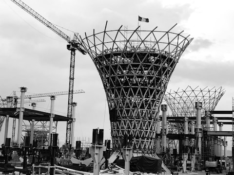 A black and white image of a construction site featuring a large, uniquely shaped structure with a lattice pattern. Several cranes can be seen in the background, along with scaffolding and various construction materials. The sky appears overcast.