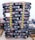 A stack of black bags labeled 'Cibau Adult 15kg', organized in a uniform manner on wooden pallets. The bags are secured with yellow and black caution tape. There is a small handwritten note placed on the stack and additional bags are visible at the side.