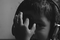 A child wearing headphones covers one eye with a hand while peering through fingers in a playful and curious manner. The photo is in black and white, casting soft shadows across the face, which highlights the youthful innocence and curiosity.
