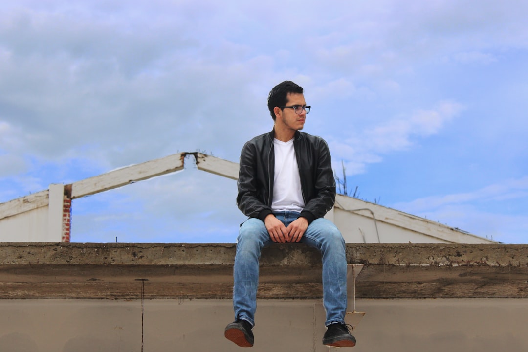 man in black jacket and blue denim jeans sitting on concrete wall