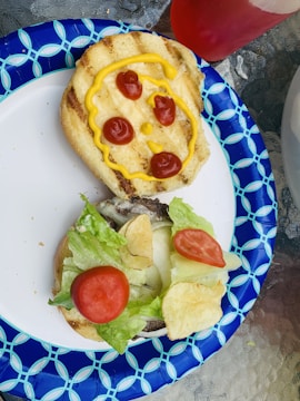 Two halves of a hamburger bun are shown on a decorative blue and white paper plate. One half has a face drawn with ketchup and mustard, featuring three ketchup spots as the eyes and mouth, and mustard for the outline. The other half contains lettuce, tomatoes, and potato chips as toppings.