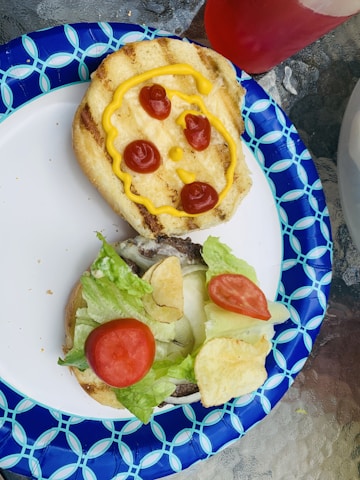 Two halves of a hamburger bun are shown on a decorative blue and white paper plate. One half has a face drawn with ketchup and mustard, featuring three ketchup spots as the eyes and mouth, and mustard for the outline. The other half contains lettuce, tomatoes, and potato chips as toppings.