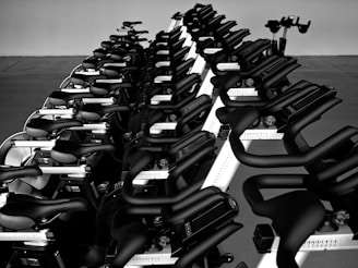 A black and white image featuring a neat row of stationary exercise bikes. The bikes are aligned closely together, emphasizing a sense of order and uniformity in a spacious, minimalistic environment.