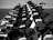 A black and white image featuring a neat row of stationary exercise bikes. The bikes are aligned closely together, emphasizing a sense of order and uniformity in a spacious, minimalistic environment.