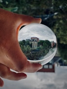 A hand holding a clear crystal ball that reflects an upside-down image of a neighborhood, including trees and brick buildings. The background is blurred, with greenery visible.