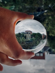 A hand holding a clear crystal ball that reflects an upside-down image of a neighborhood, including trees and brick buildings. The background is blurred, with greenery visible.