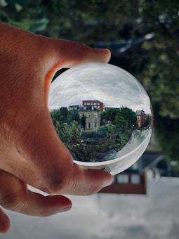 A hand holding a clear crystal ball that reflects an upside-down image of a neighborhood, including trees and brick buildings. The background is blurred, with greenery visible.
