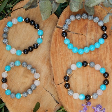 Four beaded bracelets are arranged on two pieces of rustic, natural wood. The bracelets feature round beads in blue, black, gray, and white hues. Some beads have a marbled texture, while others are solid-colored. Green foliage and small purple flowers accent the natural setting.