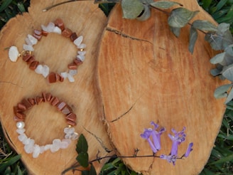 Two stone bracelets with a mix of light and dark brown hues and white stones are placed on a sliced wooden surface. A small white shell charm is attached to one bracelet. Purple flowers and green leaves surround the wood, adding a natural aesthetic.