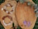 Two stone bracelets with a mix of light and dark brown hues and white stones are placed on a sliced wooden surface. A small white shell charm is attached to one bracelet. Purple flowers and green leaves surround the wood, adding a natural aesthetic.