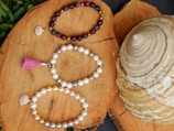 Three beaded bracelets are elegantly arranged on a natural wood surface. The top bracelet consists of dark red and gold beads with a small shell charm. The middle bracelet is made of white and light gray pearls, featuring a pink tassel. The bottom bracelet includes a mix of white, gold, and pastel-colored pearls, accompanied by a shell charm. A large, spiral-shaped seashell is visible to the right of the bracelets, adding a maritime touch.