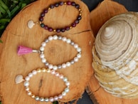 Three beaded bracelets are elegantly arranged on a natural wood surface. The top bracelet consists of dark red and gold beads with a small shell charm. The middle bracelet is made of white and light gray pearls, featuring a pink tassel. The bottom bracelet includes a mix of white, gold, and pastel-colored pearls, accompanied by a shell charm. A large, spiral-shaped seashell is visible to the right of the bracelets, adding a maritime touch.