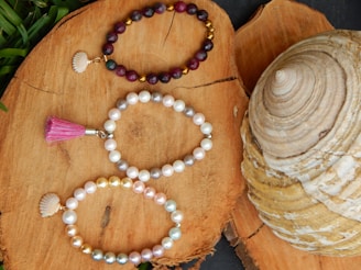 Three beaded bracelets are elegantly arranged on a natural wood surface. The top bracelet consists of dark red and gold beads with a small shell charm. The middle bracelet is made of white and light gray pearls, featuring a pink tassel. The bottom bracelet includes a mix of white, gold, and pastel-colored pearls, accompanied by a shell charm. A large, spiral-shaped seashell is visible to the right of the bracelets, adding a maritime touch.