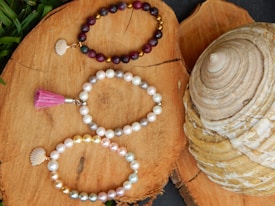 Three beaded bracelets are elegantly arranged on a natural wood surface. The top bracelet consists of dark red and gold beads with a small shell charm. The middle bracelet is made of white and light gray pearls, featuring a pink tassel. The bottom bracelet includes a mix of white, gold, and pastel-colored pearls, accompanied by a shell charm. A large, spiral-shaped seashell is visible to the right of the bracelets, adding a maritime touch.