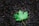 A single green maple leaf lies on a dark, textured asphalt surface. The leaf's veins and serrated edges are clearly visible, contrasting with the rough and mottled background.