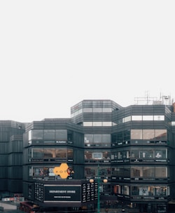 A modern, multi-story department store building with large glass windows displaying various brand names. The facade includes signs for different fashion and lifestyle brands. The structure is predominantly rectangular with geometric patterns, and the sky is overcast.