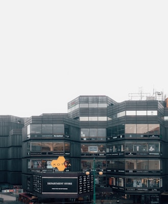 A modern, multi-story department store building with large glass windows displaying various brand names. The facade includes signs for different fashion and lifestyle brands. The structure is predominantly rectangular with geometric patterns, and the sky is overcast.