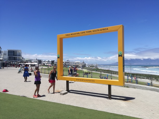 A large, rectangular yellow frame stands in an outdoor area near the coast, with people walking and taking photos around it. The frame reads 'Table Mountain, Cape Town, South Africa' and is positioned to give a scenic view. The backdrop includes the ocean, a beach, and distant mountains under a clear blue sky.