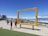 A large, rectangular yellow frame stands in an outdoor area near the coast, with people walking and taking photos around it. The frame reads 'Table Mountain, Cape Town, South Africa' and is positioned to give a scenic view. The backdrop includes the ocean, a beach, and distant mountains under a clear blue sky.