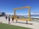 A large, rectangular yellow frame stands in an outdoor area near the coast, with people walking and taking photos around it. The frame reads 'Table Mountain, Cape Town, South Africa' and is positioned to give a scenic view. The backdrop includes the ocean, a beach, and distant mountains under a clear blue sky.