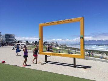 A large, rectangular yellow frame stands in an outdoor area near the coast, with people walking and taking photos around it. The frame reads 'Table Mountain, Cape Town, South Africa' and is positioned to give a scenic view. The backdrop includes the ocean, a beach, and distant mountains under a clear blue sky.
