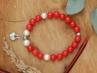 A beaded bracelet is placed on a wooden surface, featuring alternating red and white beads with a small silver heart charm attached. Surrounding the bracelet are natural elements like dried plants and green leaves, adding an organic touch to the composition.