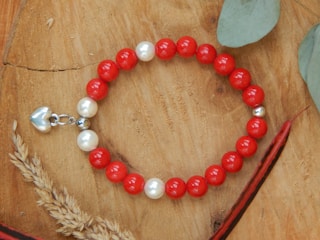 A beaded bracelet is placed on a wooden surface, featuring alternating red and white beads with a small silver heart charm attached. Surrounding the bracelet are natural elements like dried plants and green leaves, adding an organic touch to the composition.