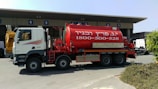 A large red tanker truck with writing on the side is parked in front of a building with numbered bays. The truck has a white cab with graphics along the side and the tanker part is prominently red with white text. The area around the truck is clear, with some bushes visible in the foreground and additional vehicles and parts of the building in the background.