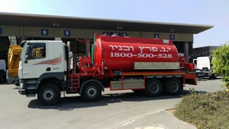 A large red tanker truck with writing on the side is parked in front of a building with numbered bays. The truck has a white cab with graphics along the side and the tanker part is prominently red with white text. The area around the truck is clear, with some bushes visible in the foreground and additional vehicles and parts of the building in the background.