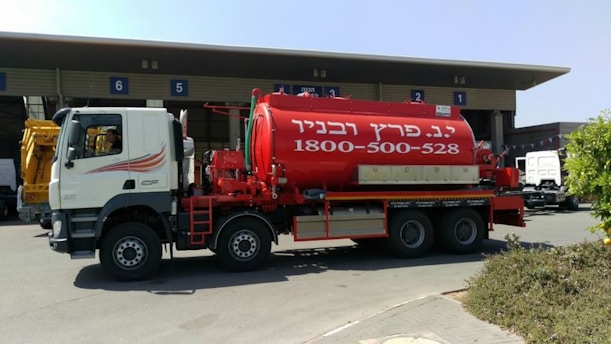 A large red tanker truck with writing on the side is parked in front of a building with numbered bays. The truck has a white cab with graphics along the side and the tanker part is prominently red with white text. The area around the truck is clear, with some bushes visible in the foreground and additional vehicles and parts of the building in the background.