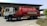 A large red tanker truck with writing on the side is parked in front of a building with numbered bays. The truck has a white cab with graphics along the side and the tanker part is prominently red with white text. The area around the truck is clear, with some bushes visible in the foreground and additional vehicles and parts of the building in the background.