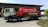 A large red tanker truck with writing on the side is parked in front of a building with numbered bays. The truck has a white cab with graphics along the side and the tanker part is prominently red with white text. The area around the truck is clear, with some bushes visible in the foreground and additional vehicles and parts of the building in the background.